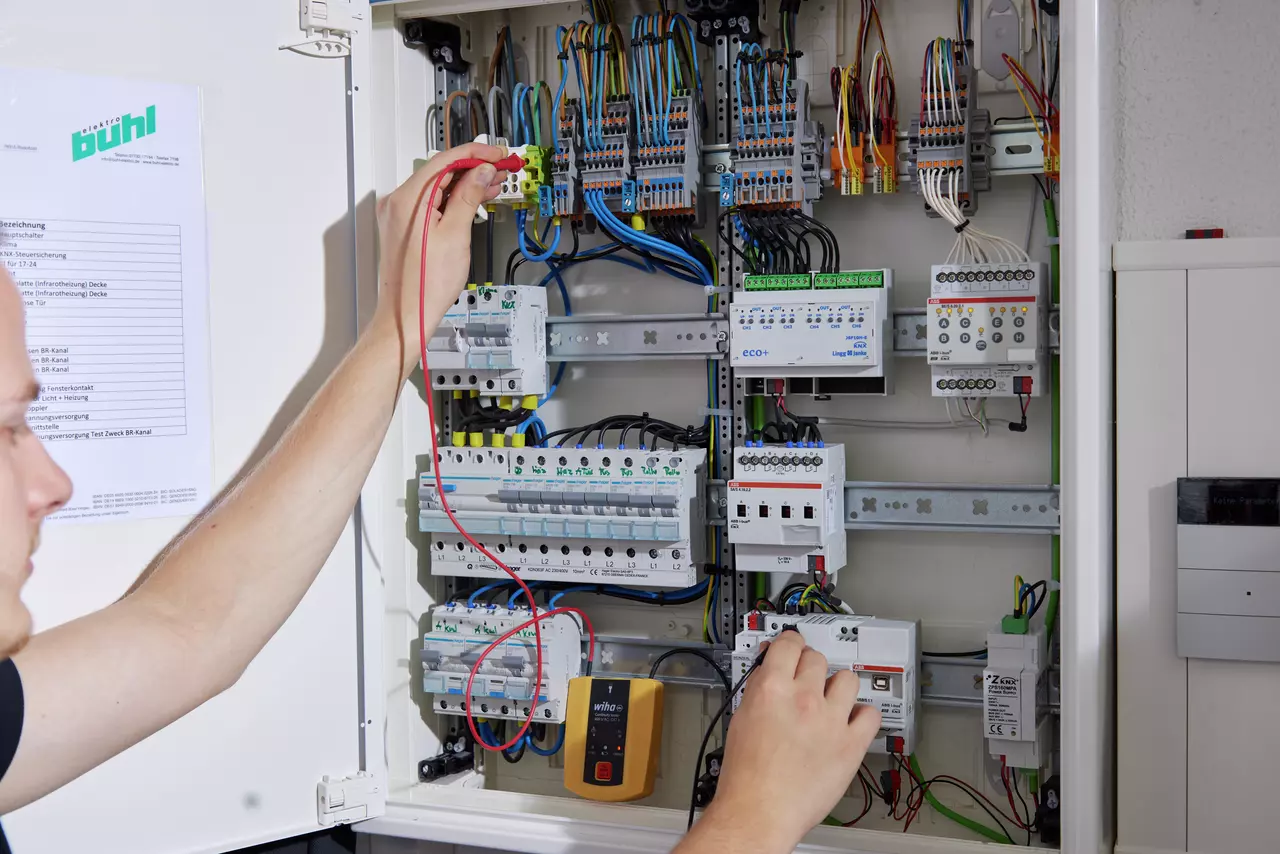 Contrôle de continuité dans une armoire électrique ouverte : électricien appliquant la pointe de touche rouge sur un bornier, testeur Wiha 45222 posé dans l’armoire devant les disjoncteurs